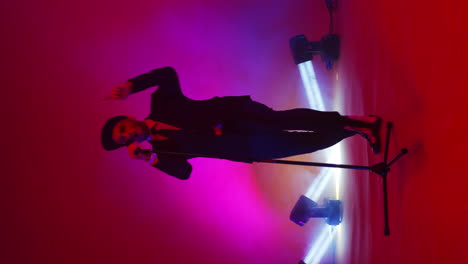 Vertical-video.-A-male-singer-with-a-microphone-stand-dances-and-sings-in-the-neon-light-in-the-Studio.-An-energetic-and-expressive-singer
